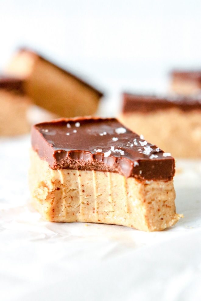 This is a side view of a chocolate almond butter bar with a bite taken out of it. The bar sits on a white piece of parchment paper and is sprinkled with salt. More bars are blurred in the background.