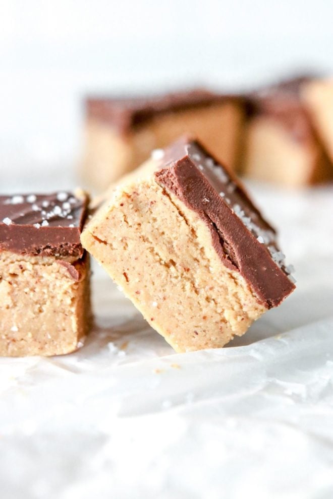 This is a side image of a chocolate almond butter bar leaning against another bar. The bars sit on a white piece of parchment paper with more bars blurred in the backrgound.
