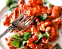This is a close up of a fork piercing a cauliflower floret coated in bbq sauce. A white plate with more bbq cauliflower is blurred in the background. The cauliflower is topped with fresh cilantro and the plate sits on a white counter.
