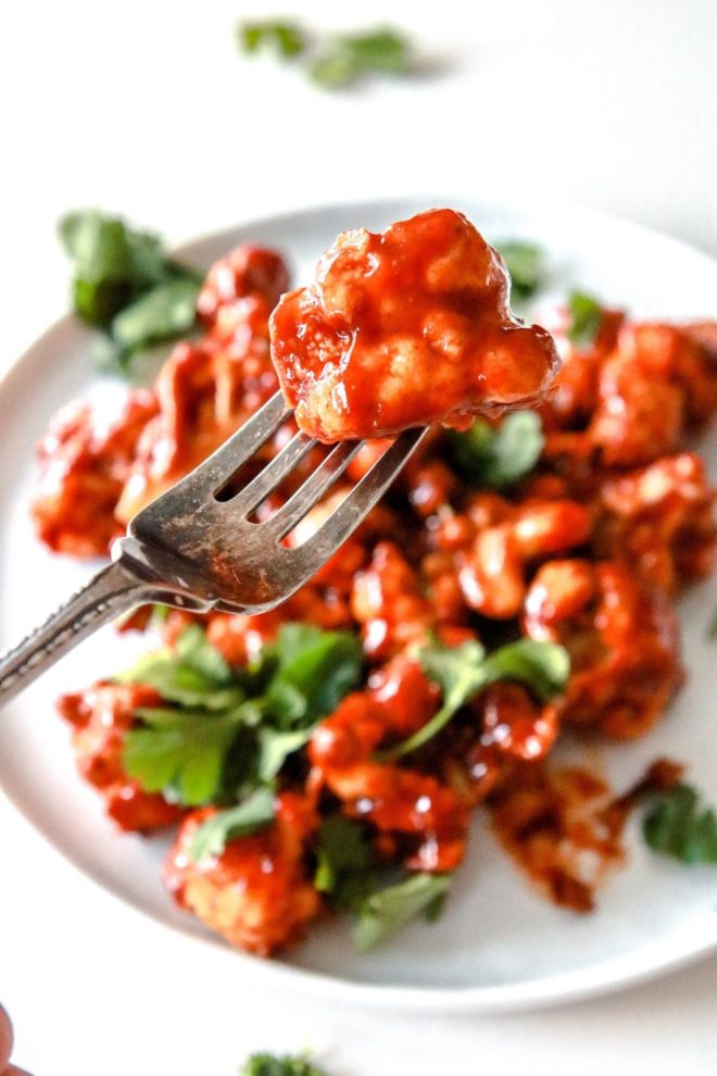 This is a close up of a fork piercing a cauliflower floret coated in bbq sauce. A white plate with more bbq cauliflower is blurred in the background. The cauliflower is topped with fresh cilantro and the plate sits on a white counter.