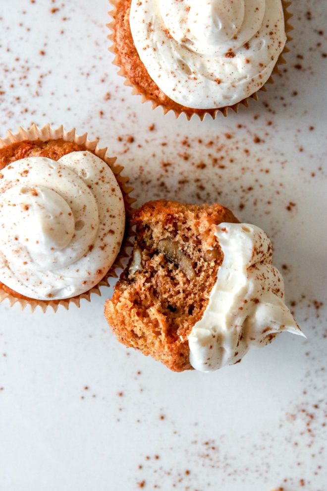 This is an overhead image of a carrot cake cupcake with cream cheese frosting. Cinnamon is sprinkled on top of the cupcakes and on the white counter around the cupcakes. One cupcake has a bite taken out and is leaning on its side against another cupcake.