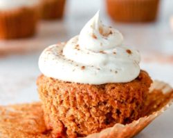 This is a side view of a carrot cake cupcake topped with cream cheese frosting and sprinkled with cinnamon. The cupcake sits on a white counter with more cupcakes blurred in the background. The paper cupcake liner is peeled off the cupcake revealing the fluffy cake.