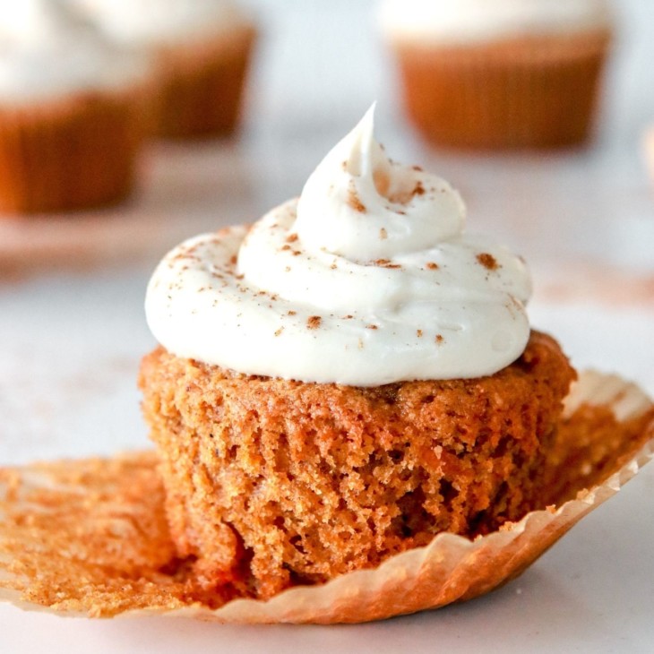 This is a side view of a carrot cake cupcake topped with cream cheese frosting and sprinkled with cinnamon. The cupcake sits on a white counter with more cupcakes blurred in the background. The paper cupcake liner is peeled off the cupcake revealing the fluffy cake.