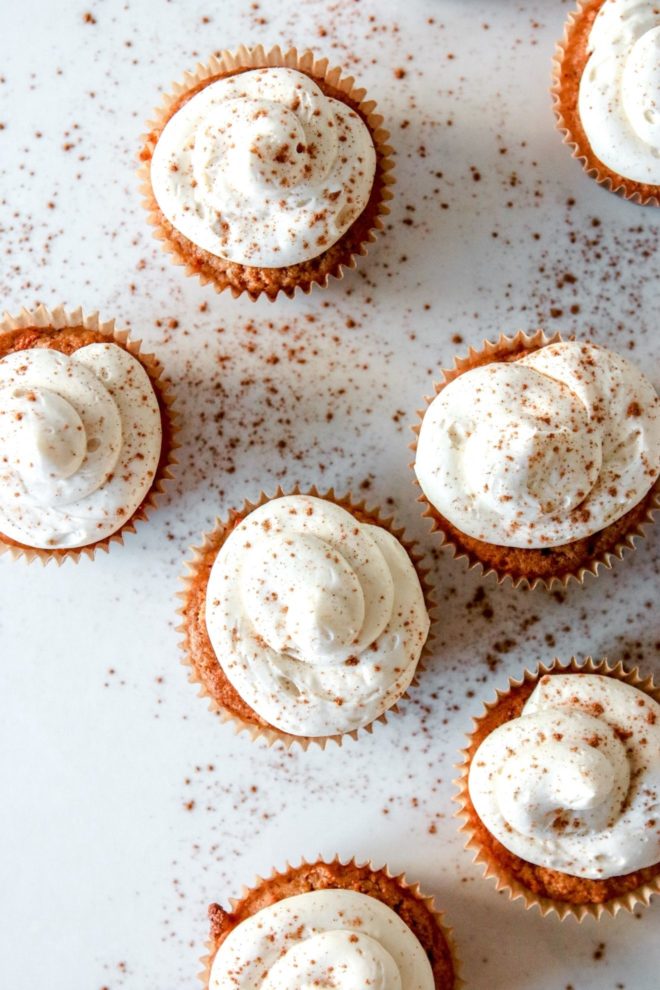This is an overhead image of cupcakes with a vanilla frosting. The cupcakes are sprinkled with cinnamon and are sitting on a white counter.