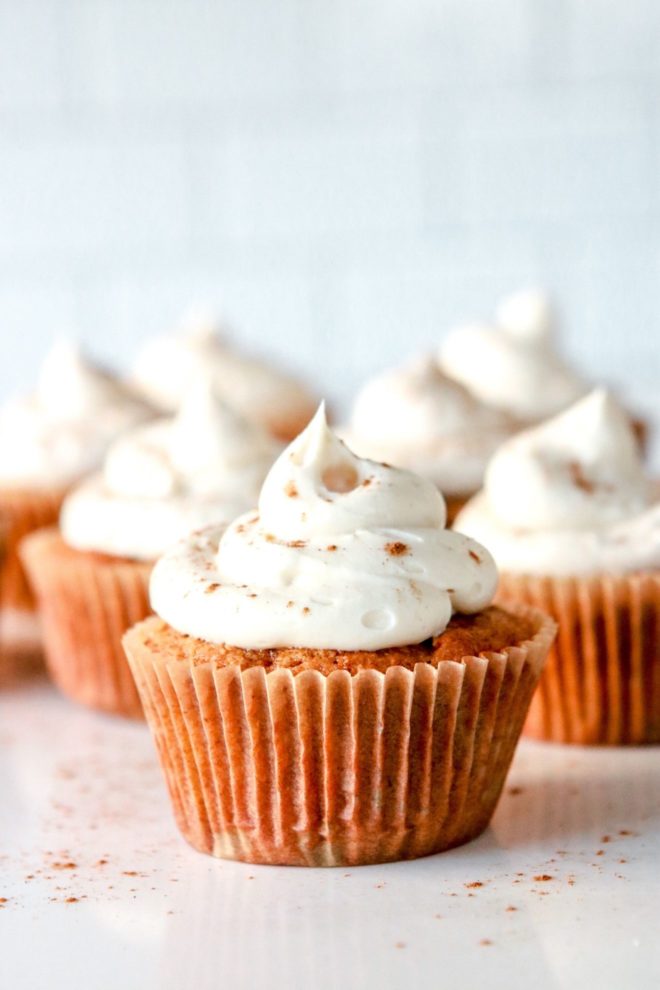 This is a side view of a cupcakes with a white frosting. The cupcakes are sprinkled with cinnamon and sitting on a white counter. One cupcake is close and in focus while the rest of the cupcakes are blurred in the background.