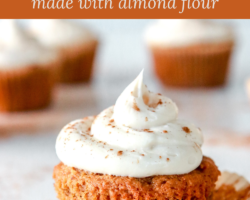 This is a side view of a carrot cake cupcake topped with cream cheese frosting and sprinkled with cinnamon. The cupcake sits on a white counter with more cupcakes blurred in the background. The paper cupcake liner is peeled off the cupcake revealing the fluffy cake. Text overlay reads "carrot cake cupcakes made with almond flour."