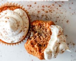 This is an overhead image of a carrot cake cupcake with cream cheese frosting. Cinnamon is sprinkled on top of the cupcakes and on the white counter around the cupcakes. One cupcake has a bite taken out and is leaning on its side against another cupcake. Text overlay reads "carrot cake cupcakes made with almond flour."