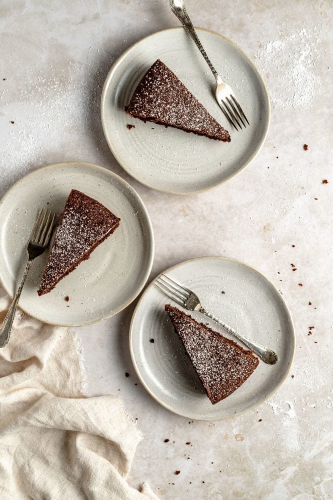This is an overhead image of a small plates with a slices of chocolate olive oil cake. A fork is laying next to the cake on the plates. The chocolate cake is sprinkled with powdered sugar.