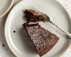 This is an overhead image of a small plate with a slice of chocolate olive oil cake. A fork is cutting off a bite of cake. The chocolate cake is sprinkled with powdered sugar.