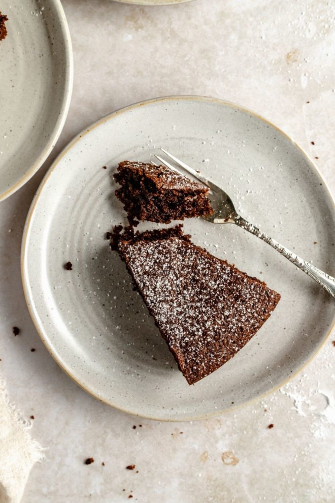 This is an overhead image of a small plate with a slice of chocolate olive oil cake. A fork is cutting off a bite of cake. The chocolate cake is sprinkled with powdered sugar.