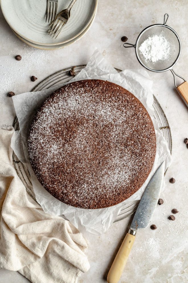 This is an overhead image of chocolate olive oil cake. The cake sits on a piece of white parchment paper on a cooling rack. The cake is sprinkled with powdered sugar. A knife, chocolate chips, a tan tea towel, a sifter with powdered sugar, and small plated with forks on top are on the counter around the cake.