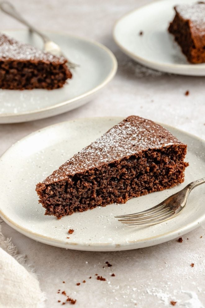 This is a side view of a slice of chocolate olive oil cake on a plate with a fork. The cake sits on a light color surface with more slices of cake blurred in the background.