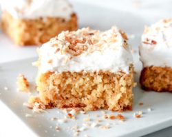 This is a side view of a slice of coconut cake topped with vanilla frosting and toasted coconut shreds. The cake sits on a small white rectangular plate with more slices of cake blurred in the background.