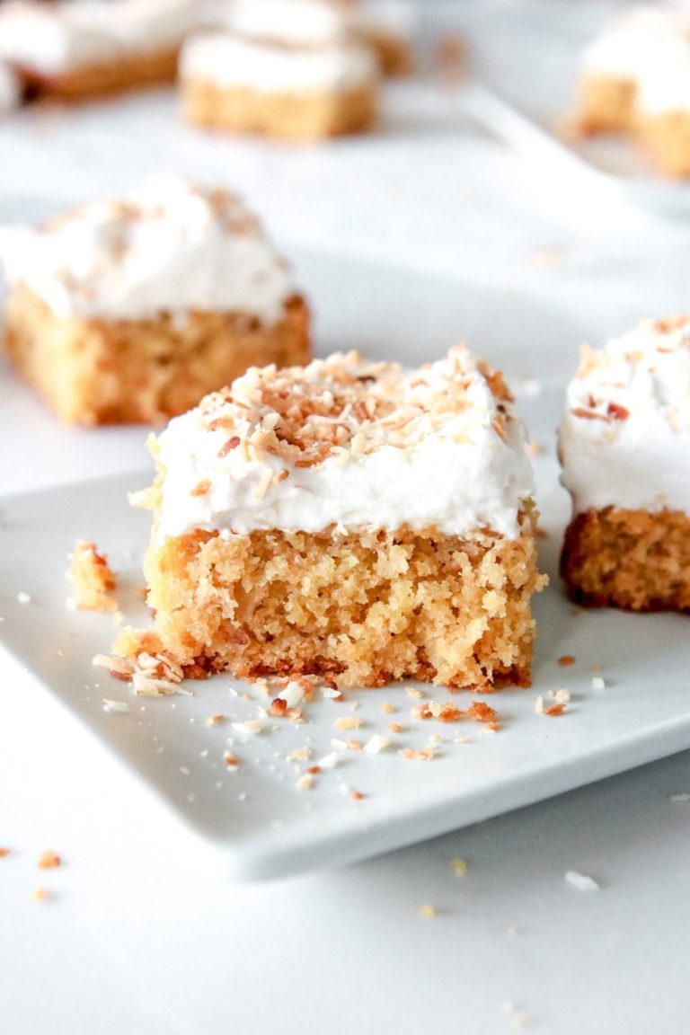 This is a side view of a slice of coconut cake topped with vanilla frosting and toasted coconut shreds. The cake sits on a small white rectangular plate with more slices of cake blurred in the background.