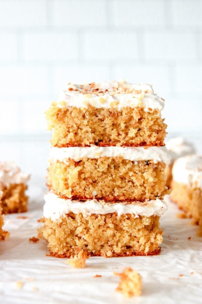 This is a stack of three pieces of cake sitting on a white piece of parchment paper. More pieces of cake are blurred in the background and a white tile background is in the distance behind that stack of cake.