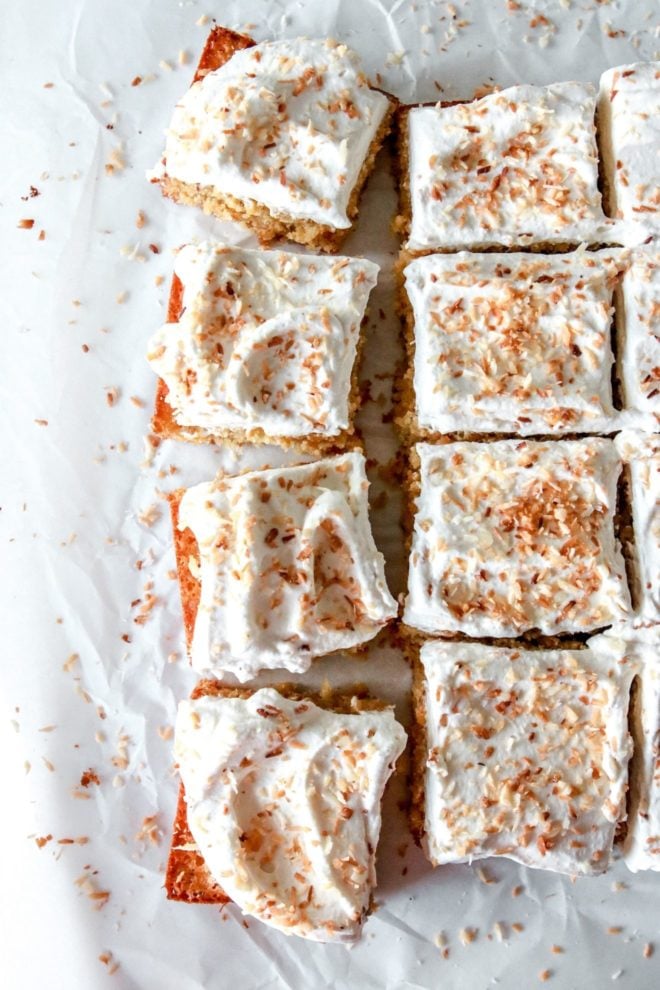 This is an overhead image of coconut cake cut in squares. The cake sits on a white piece of parchment paper and are separated and pulled away from the other pieces. The cake is topped with white frosting and toasted coconut.