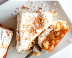 This is an overhead image of a square piece of cake on a small white rectangular plate on a white counter. A fork is cutting off a piece of the cake and laying against the side of the plate. Text overlay reads "gluten free coconut cake."