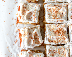 This is an overhead image of coconut cake cut in squares. The cake sits on a white piece of parchment paper and are separated and pulled away from the other pieces. The cake is topped with white frosting and toasted coconut. Text overlay reads "toasted coconut cake."