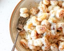 This is an overhead image of a bowl with gnocchi and gorgonzola sauce. A fork lays against the side of the bowl piercing a few gnocchis. The bowl sits on a white counter with some ground pepper. A blue tea towel is in the top left corner of the image.