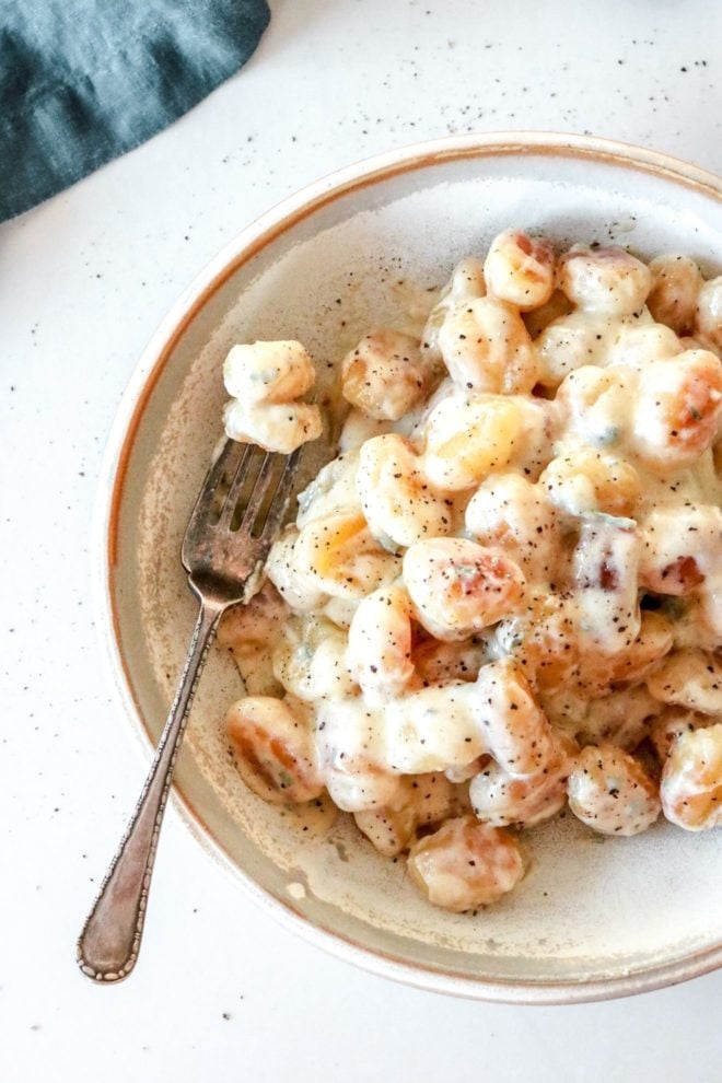 This is an overhead image of a bowl with gnocchi and gorgonzola sauce. A fork lays against the side of the bowl piercing a few gnocchis. The bowl sits on a white counter with some ground pepper. A blue tea towel is in the top left corner of the image.