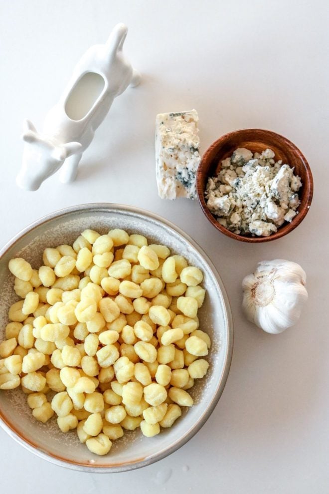 This is an overhead view of ingredients on a white counter. The ingredients are gnocchi, a garlic head, crumbled gorgonzola, and a small cow shape pitcher filled with cream.