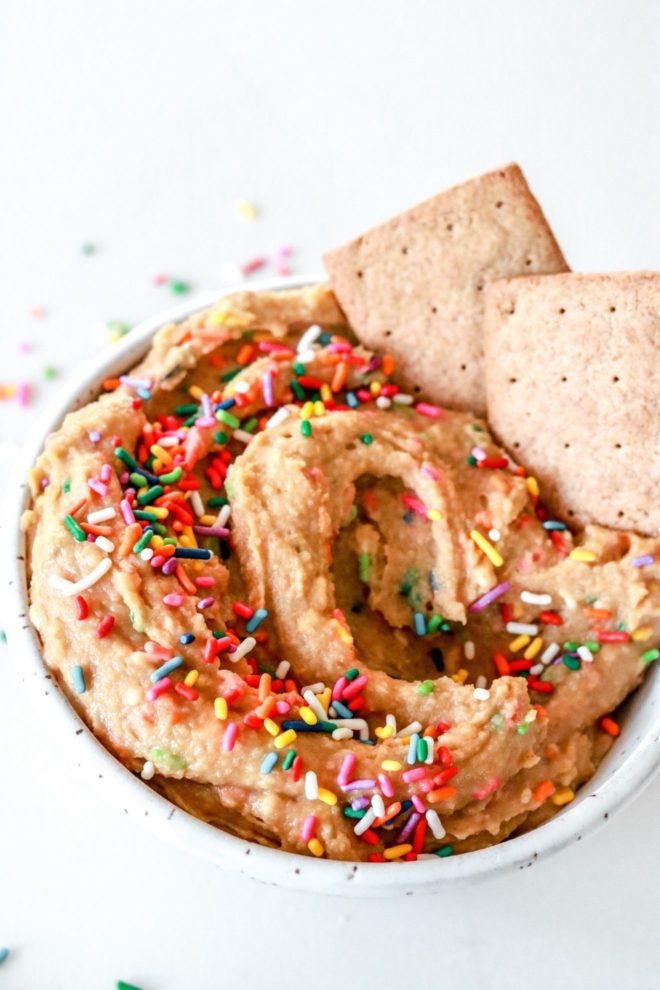 This is a side view of funfetti dip with rainbow sprinkles. Graham crackers are dipped in the bowl on the right hand side. The bowl sits on a white counter with more rainbow sprinkles scattered around the bowl.