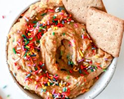 This is an overhead image of funfetti dip with rainbow sprinkles. Graham crackers are dipped in the bowl on the right hand side. The bowl sits on a white counter with more rainbow sprinkles scattered around the bowl.