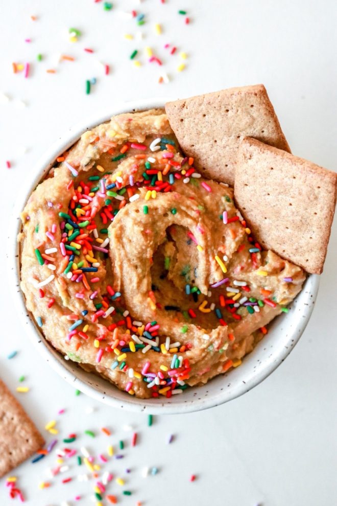This is an overhead image of funfetti dip with rainbow sprinkles. Graham crackers are dipped in the bowl on the right hand side. The bowl sits on a white counter with more rainbow sprinkles scattered around the bowl.