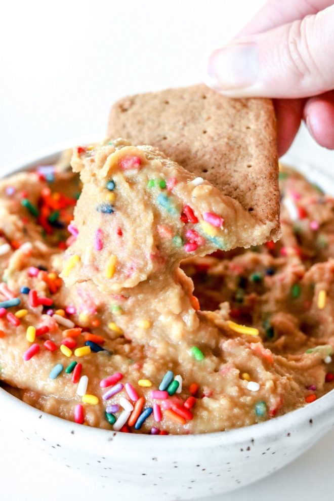 A hand is dipping a graham cracker into cake batter dip with rainbow sprinkles. The dip is in a white speckled bowl sitting on a white counter.
