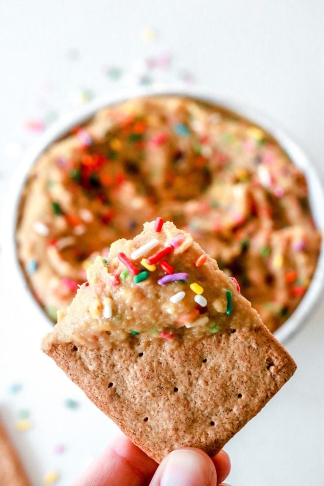 A hand is holding a graham cracker dipped in cake batter dip with rainbow sprinkles. A white bowl filled with funfetti dip is blurred on a white counter on the background.