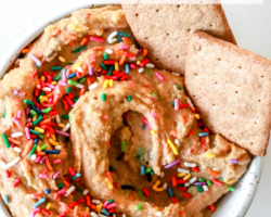This is an overhead image of funfetti dip with rainbow sprinkles. Graham crackers are dipped in the bowl on the right hand side. The bowl sits on a white counter with more rainbow sprinkles scattered around the bowl. Text overlay reads "healthy cake batter funfetti dip."