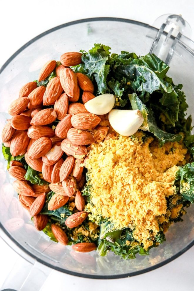 This image looks into a food processor filled with kale, almonds, garlic, and nutritional yeast. The food processor sits on a white counter.