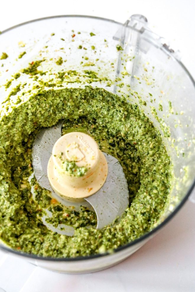 This images looks into a food proecessor filled with pesto. The jar sits on a white counter.