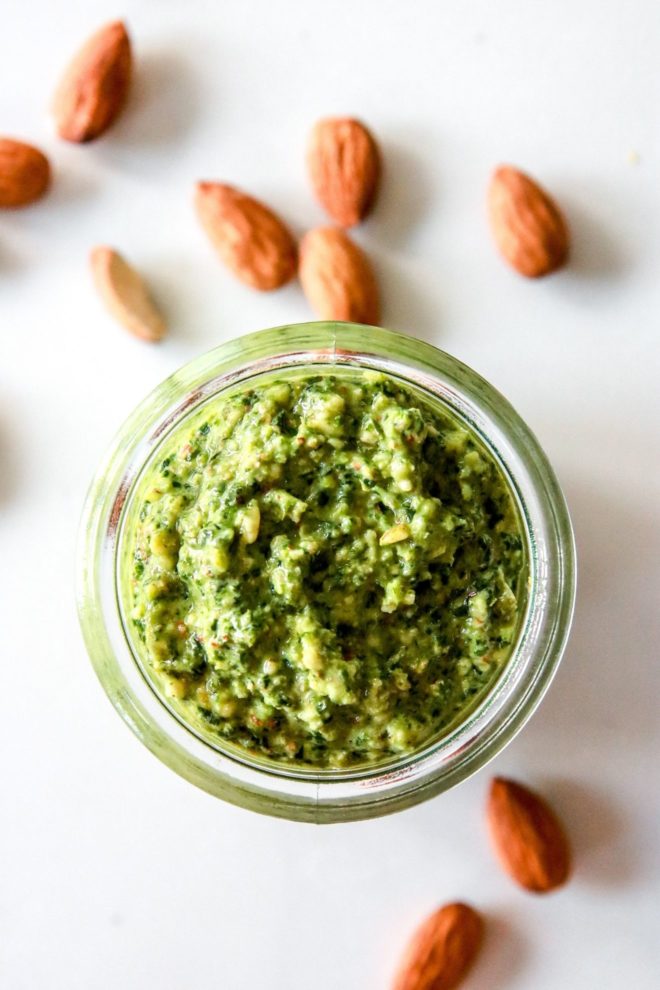 This images looks into a glass jar filled with pesto. The jar sits on a white counter scattered with almonds.