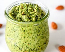 This images looks into a glass jar filled with pesto. The jar sits on a white counter scattered with almonds.