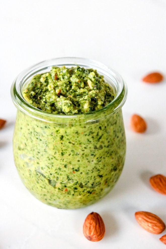 This images looks into a glass jar filled with green kale almond pesto sauce. The jar sits on a white counter scattered with almonds.