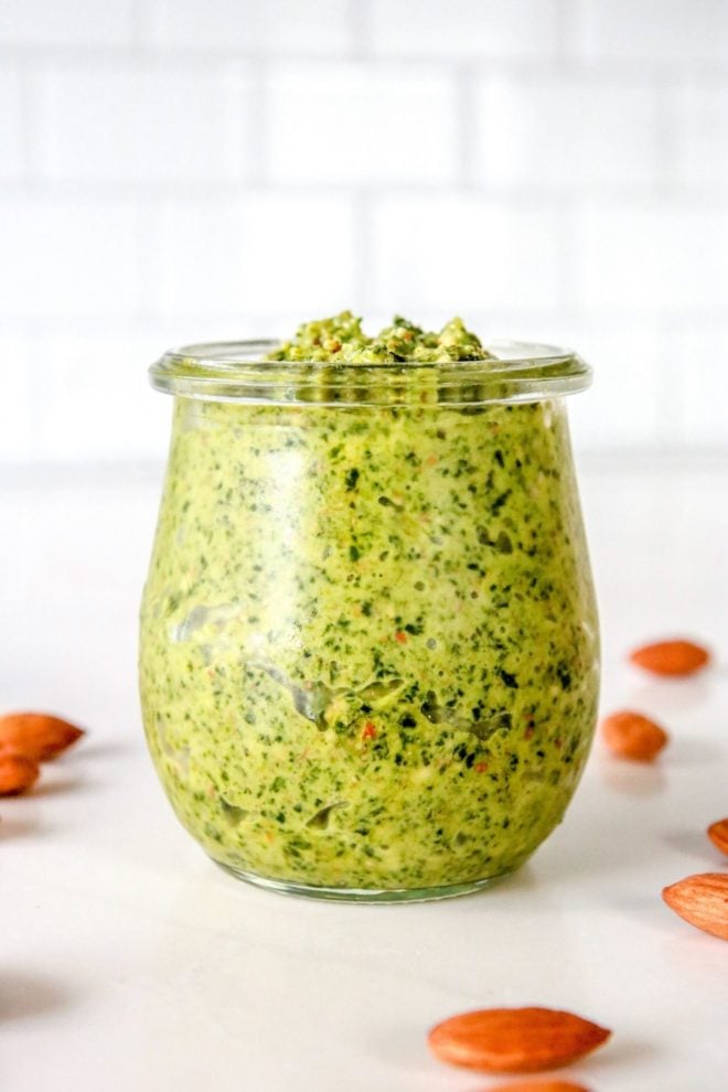This is a side view of a small glass jar filled with green kale almond pesto sauce. The jar sits on a white counter surround with almonds and a white tile background.