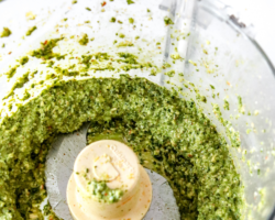 This images looks into a food processor filled with pesto. The jar sits on a white counter. Text overlay reads "vegan almond kale pesto."