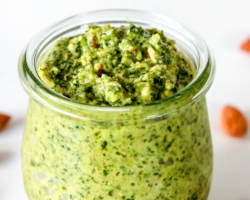 This images looks into a glass jar filled with pesto. The jar sits on a white counter scattered with almonds. Text overlay reads "vegan almond kale pesto."