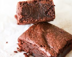 A fudgey chocolate brownie leans against another brownie. The brownies sit on a white piece of parchment paper and are sprinkled with cocoa powder.