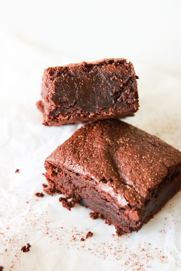 A fudgey chocolate brownie leans against another brownie. The brownies sit on a white piece of parchment paper and are sprinkled with cocoa powder.