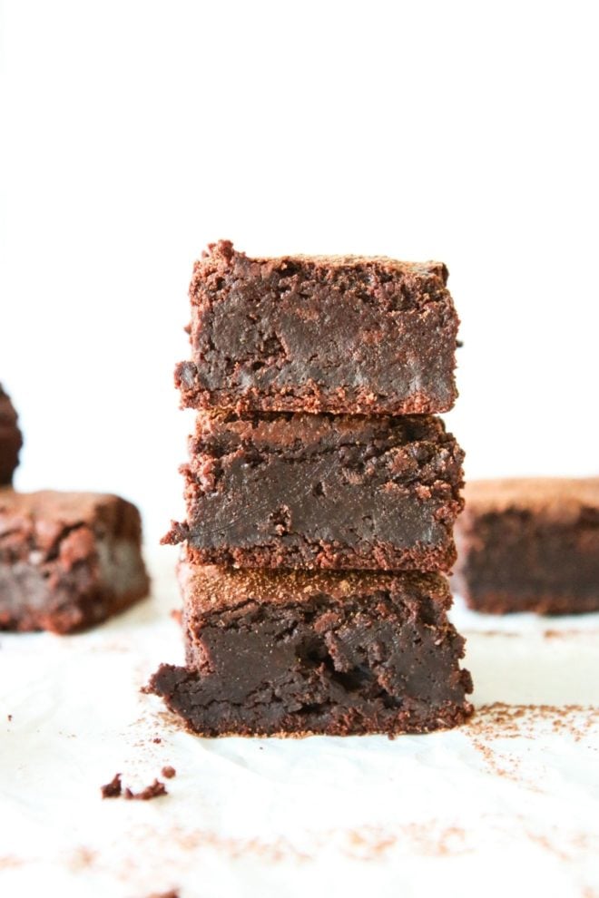 This is a stack of three fudgey brownies. The brownies sit on a white piece of parchment paper and a white background. More brownies are blurred in the background.