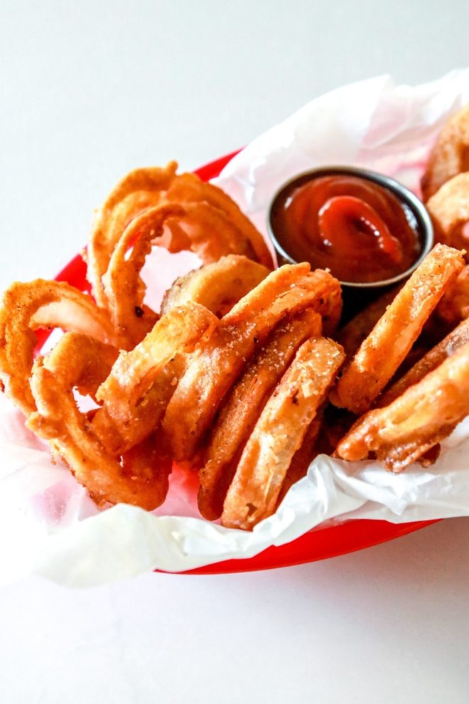 This is a side view of onion rings in a red basket with parchment paper. A small silver cup filled with ketchup is also in the basket.