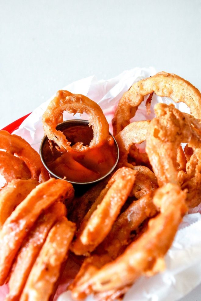 This is a side view of battered and fried onions in a red basket with parchment paper. One onion is being dipped into a small bowl of ketchup.