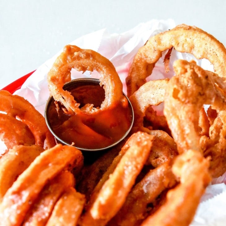 This is a side view of onion rings in a red basket with parchment paper. One onion rings is being dipped into a small bowl of ketchup.
