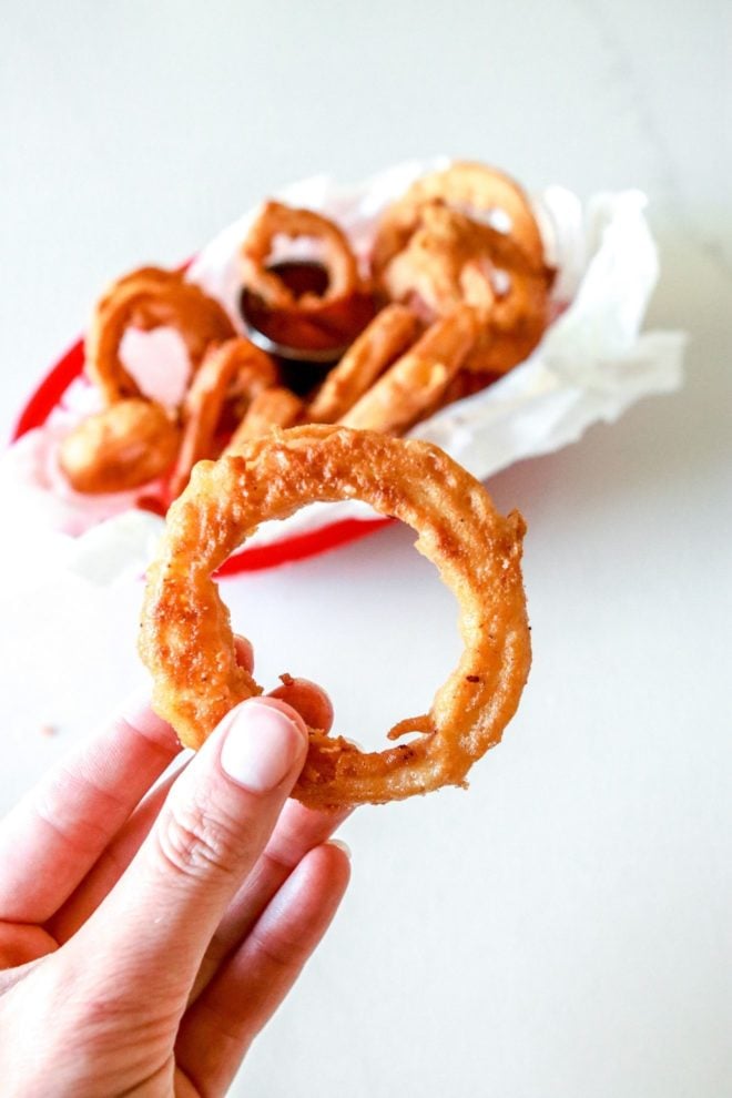 A hand is holding a fried onion ring. A red basket with parchment paper is blurred in the background and filled with more onion rings.