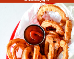 This is an overhead view of onion rings in a red basket with parchment paper. A small silver cup filled with ketchup is also in the basket. Text overlay reads "gluten free crispy onion rings"