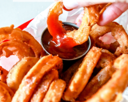 A hand is dipping an onion ring into a small silver cup of ketchup. The basket is lined with parchment paper and filled with more onion rings. Text overlay reads "gluten free crispy onion rings."
