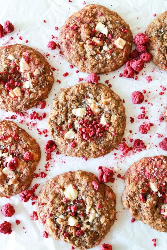 This is an overhead image of cookies with freeze dried raspberry pieces and white chocolate. The cookies lie on a piece of parchment paper. More freeze dried raspberries are on top and around the cookies.