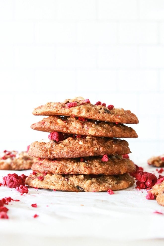 This is a side view of a stack of cookies. The stack sits on a white piece of parchment paper and a white background. Freeze dried raspberries are around the cookies.